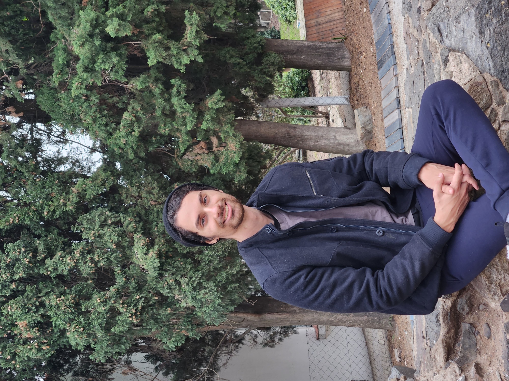 Photo of Guilherme Ramos, a product designer, smiling while seated outdoors in a wooded area, wearing a dark coat and a beanie.