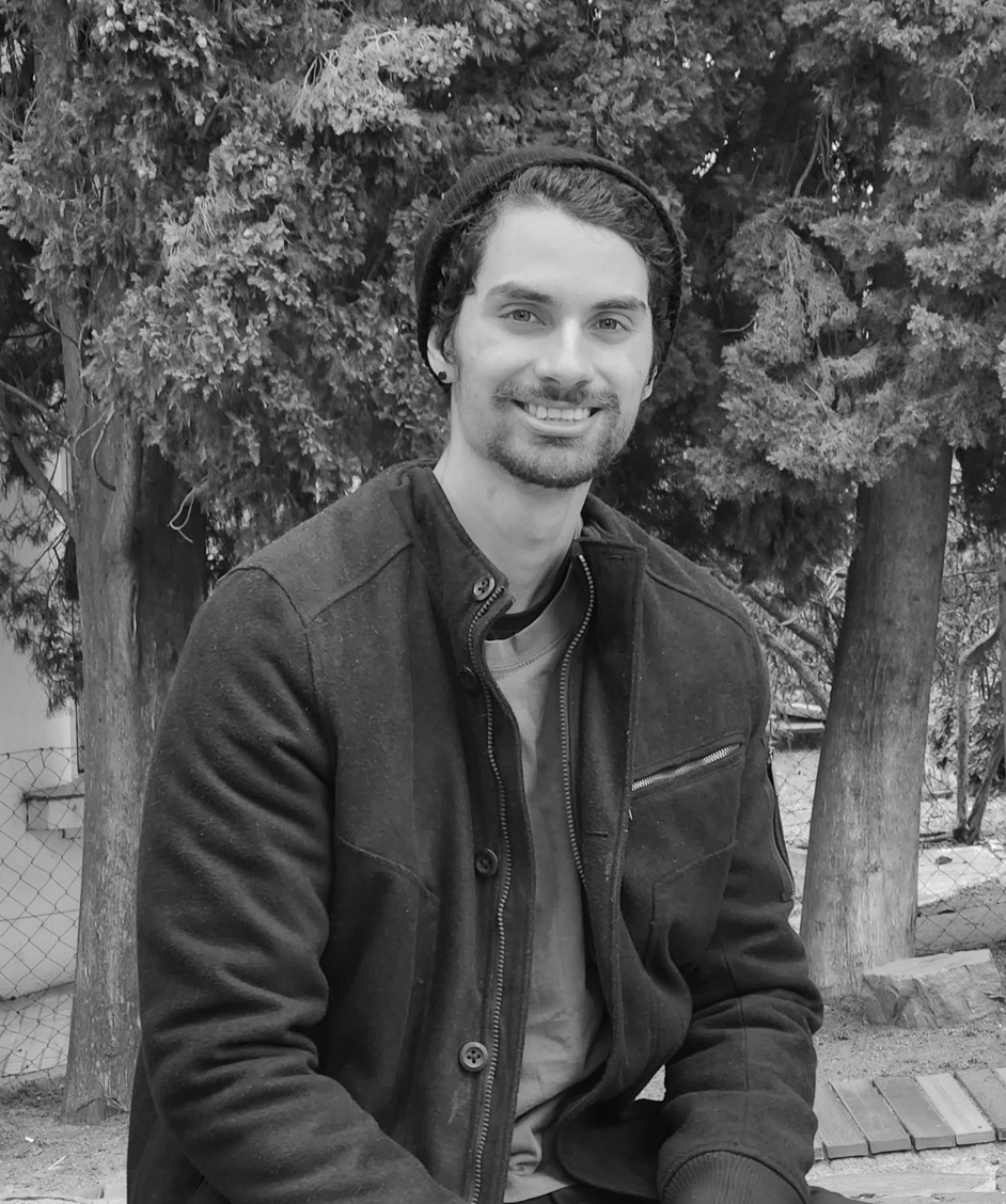 Photo of Guilherme Ramos, a product designer, smiling while seated outdoors in a wooded area, wearing a dark coat and a beanie.