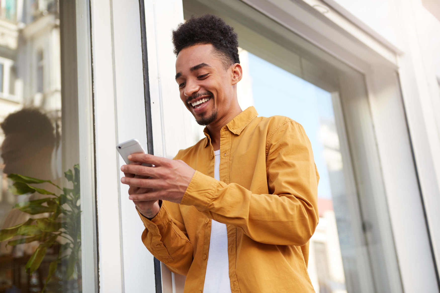 Photo of a dark-skinned happy young man in a yellow shirt walking down the street, holding the phone, talking to his girlfriend, and smiling, looking cheerful.